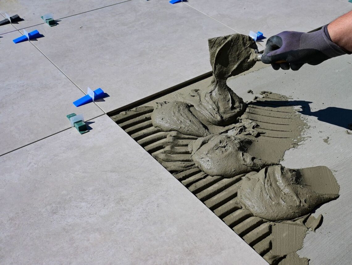 Worker pours cement grout beneath aligned floor tiles using a trowel