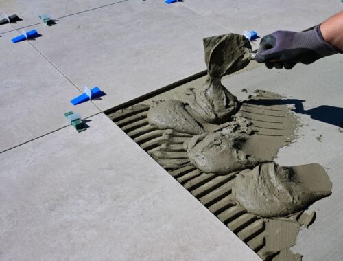 Worker pours cement grout beneath aligned floor tiles using a trowel